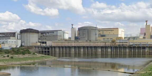 Industrial facility with large storage tanks and a dam along a calm river under a partly cloudy sky.