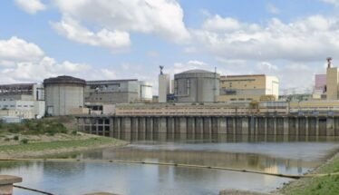 Industrial facility with large storage tanks and a dam along a calm river under a partly cloudy sky.