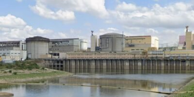 Industrial facility with large storage tanks and a dam along a calm river under a partly cloudy sky.