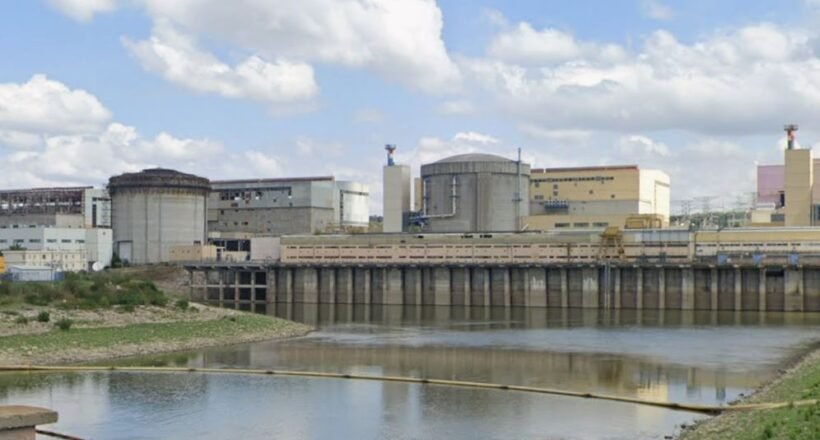 Industrial facility with large storage tanks and a dam along a calm river under a partly cloudy sky.