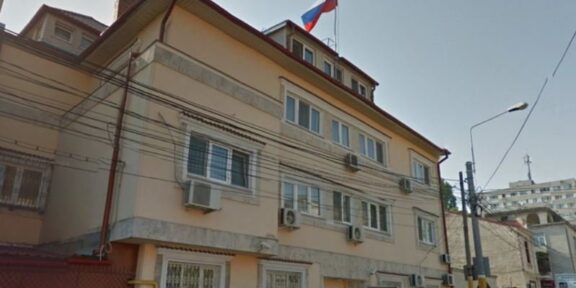Beige three-story building with window air conditioners, a Russian flag flying from the roof, and a narrow street with a streetlight nearby.