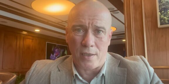 Close-up of a bald man in a gray suit speaking to the camera in a wood-paneled room.