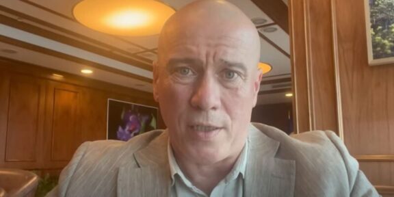 Close-up of a bald man in a gray suit speaking to the camera in a wood-paneled room.