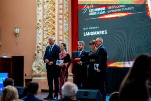 Group of formally dressed award recipients posing on a stage, holding plaques, during a ceremony with ornate decor and a large screen behind them.]