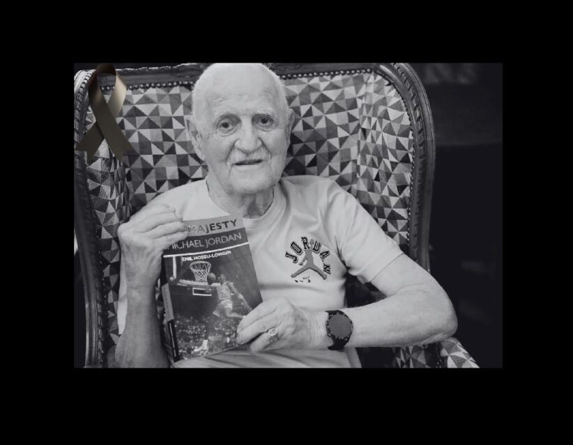 Older man with a white T-shirt sits in a patterned chair, holding a Michael Jordan basketball book and wearing a watch; a black ribbon is pinned to the chair in the background.