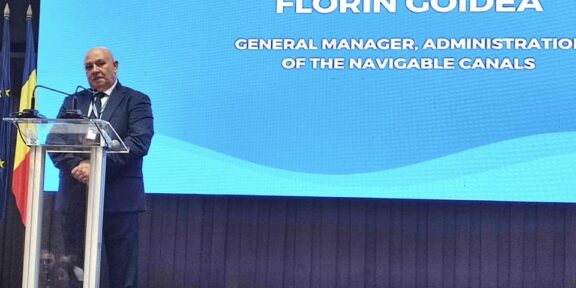 Man in a dark suit giving a speech at a clear podium on a stage, with a large blue presentation screen behind him and a Romanian flag visible on the left