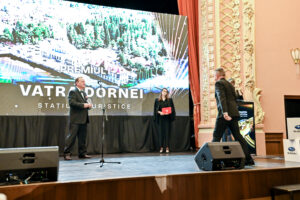 Man in a suit on a stage speaks at a microphone while holding an award; a woman in black stands beside him holding a red booklet, with a large screen backdrop and ornate gold theater walls to the right.