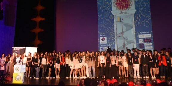 Large group of young people on a theater stage posing for a group photo during an event, with a tall blue graphic poster projected on a screen behind them and red balloons in the foreground.
