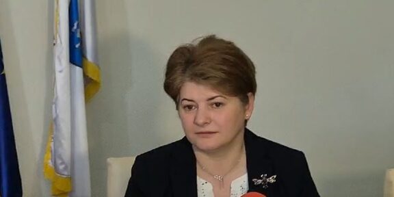 Woman in a dark blazer speaks into a red microphone during an interview, with flags in the background.