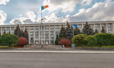 Front of a large government building with steps, flagpoles, and manicured trees in front of it in an urban plaza.
