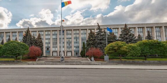 Front of a large government building with steps, flagpoles, and manicured trees in front of it in an urban plaza.