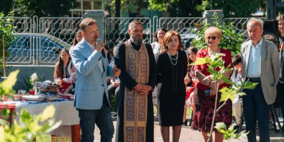 Man in light blue blazer speaks into a microphone at an outdoor ceremony, surrounded by adults in formal attire and traditional clothing.