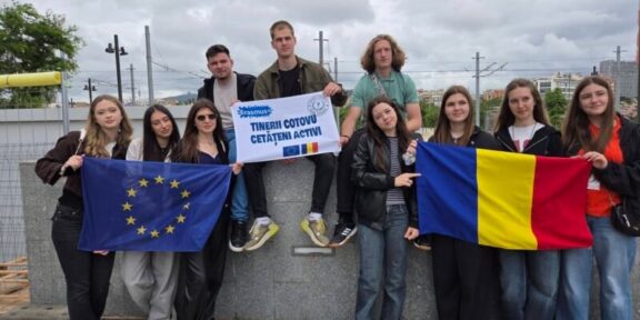 Group of diverse young students on a rooftop, holding the EU flag and the Romanian flag with an Erasmus banner saying active citizens.