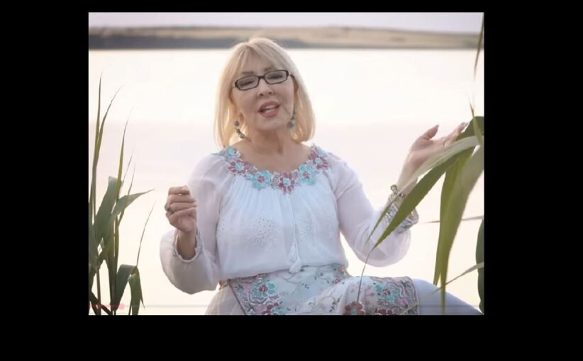 Older woman with blonde hair and glasses sits by a lake, wearing a white embroidered blouse and turquoise accessories, speaking or singing to the camera.