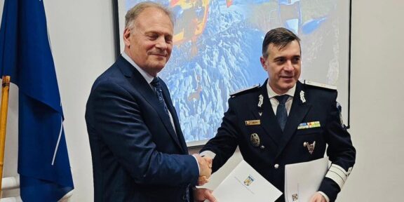 Two men shake hands in a formal room; one in a suit and the other in a military uniform with medals, holding documents behind them with a projector screen the background showing helicopters and a flag on the left side.