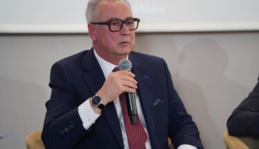 Man in a navy suit and red tie speaks into a handheld microphone at a panel discussion.