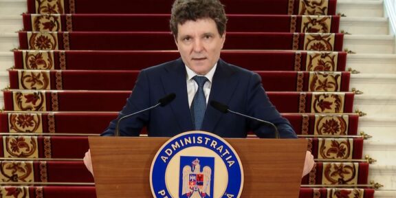 A man in a dark suit stands behind a wooden podium with a blue circular seal reading Administratia Prezidentială, speaking into two microphones, with red-carpeted stairs in the background.