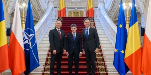 Three men in dark suits pose on a red-carpeted grand staircase, flanked by national flags including Romania, Poland, NATO, and the EU flag in a gilded hall.
