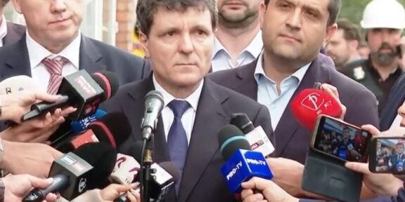 Man in a dark suit giving a press statement, surrounded by reporters and microphones outdoors.