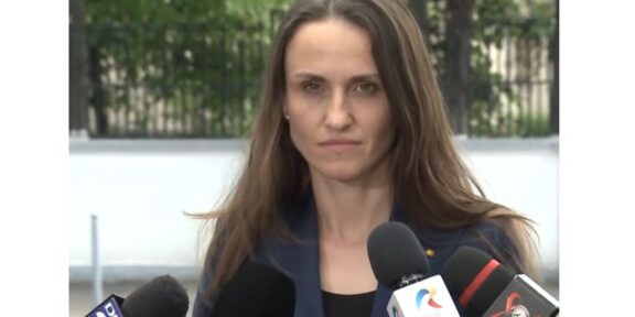 Woman in a navy blazer speaks to reporters at an outdoor press conference, multiple microphones in front of her.