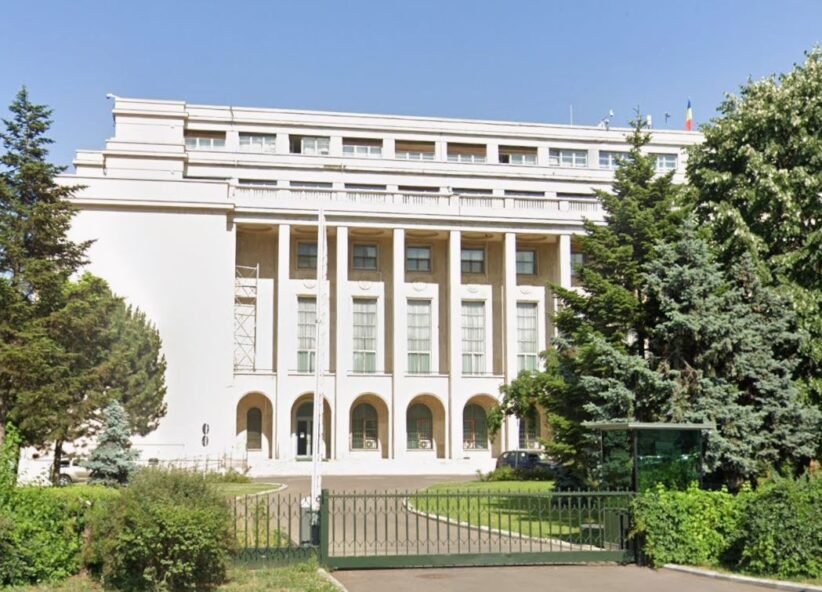 White neoclassical building with tall columns, arches at ground level, gated lawn, and surrounding trees.