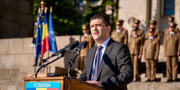 Man in a dark suit delivering a speech at a wooden podium labeled ROMANIA, with flags and soldiers in ceremonial uniforms behind him.