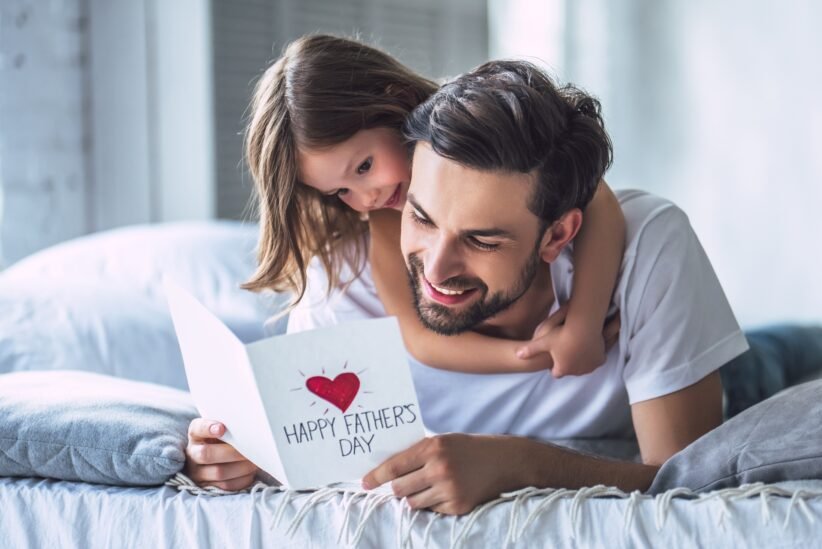 Father and daughter cuddling on a bed, smiling at a card that reads 'Happy Father's Day' with a red heart.