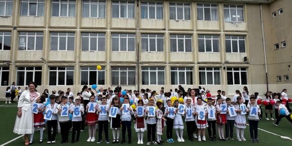 Group of primary school children in white shirts and traditional outfits, standing on a field in front of a beige school building, each holding a card with a letter to form a message.