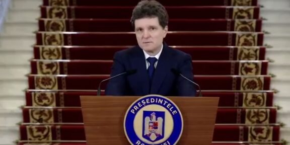 Man in a dark suit delivering a formal speech at a wooden podium with a round blue-and-white seal, in front of a red-carpeted staircase backdrop.