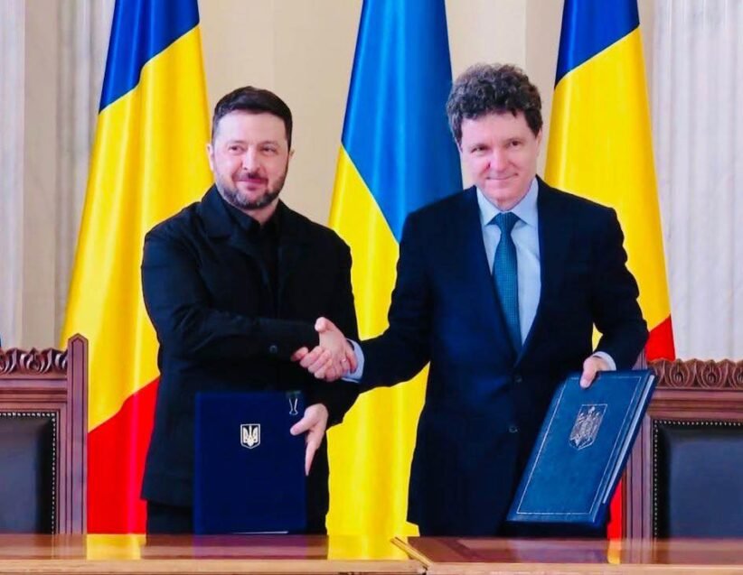 Two men in formal attire shake hands over a table, each holding a blue folder with a crest, Ukrainian flags in the background.