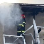 Firefighter in protective gear and red helmet spraying water as smoke pours from a fire-damaged building exterior.