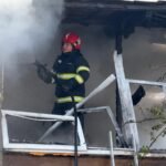 Firefighter in red helmet douses a burning building from a second-floor balcony amid smoke and charred debris.