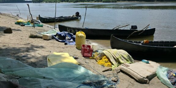 A polluted riverside beach with several wooden boats pulled up on shore and scattered debris, including nets, tarps, and plastic containers. Trees across the water in the background.