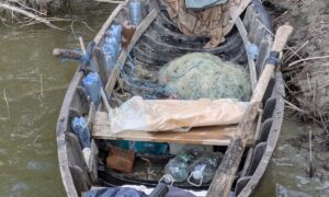 A weathered wooden boat cluttered with plastic bottles, fishing nets, and bags, stranded in murky water near a muddy shoreline.
