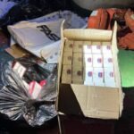 Open cardboard box filled with many small white boxes with blue and red labels, surrounded by bags of trash and a red metal drum on a dirty floor.