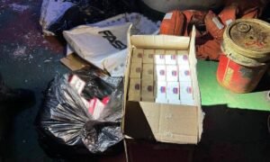Open cardboard box filled with many small white boxes with blue and red labels, surrounded by bags of trash and a red metal drum on a dirty floor.
