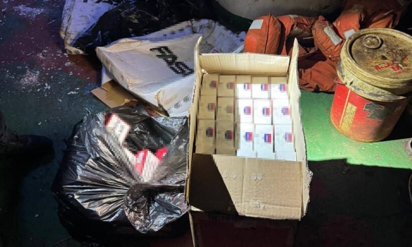 Open cardboard box filled with many small white boxes with blue and red labels, surrounded by bags of trash and a red metal drum on a dirty floor.