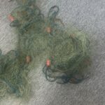 Tangled green fishing net with blue lines and orange floats on a gray carpet. A plastic bag sits nearby for scale.