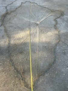 Large fishing net spread flat on a concrete surface, with a central knot and a yellow measuring tape laid along its length for scale.