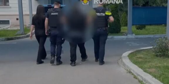 Police officers escorting a blurred person down a street near a building entrance with a blue canopy; 'Politia Romana' emblem visible