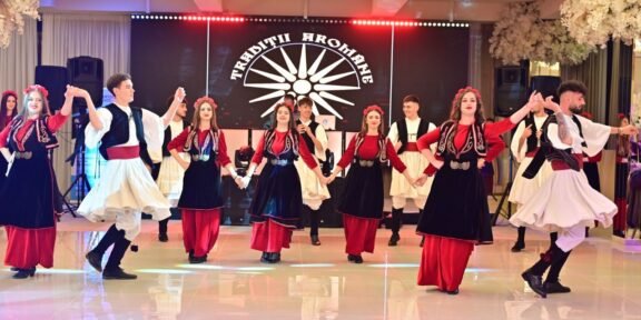 Dancers in red, black, and white traditional outfits perform a coordinated folk dance on a stage with a sunburst backdrop.