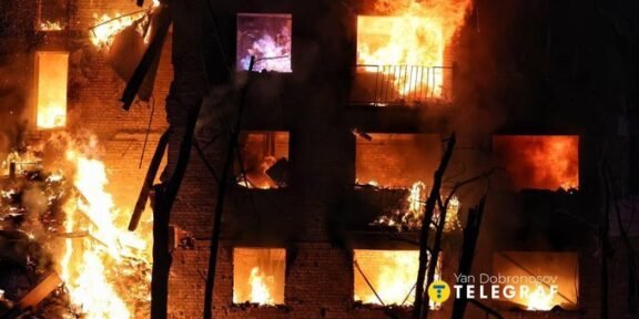 A brick building is engulfed in bright orange flames at night, with multiple windows burning and smoke filling the scene.