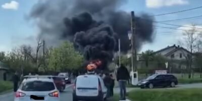 Car on a two-lane road is on fire, sending a large plume of black smoke while bystanders watch and other cars stop nearby on a residential street.