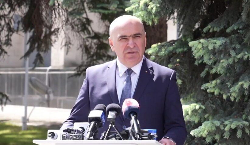 Man in a navy suit delivering a press conference outdoors, with multiple microphones on a podium in front of him.