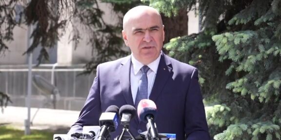 Man in a navy suit delivering a press conference outdoors, with multiple microphones on a podium in front of him.