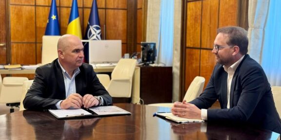 Two men in suits sit at a large polished conference table, papers and notebooks in front, with flags and wood paneling in a formal office behind them.