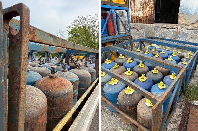 Rows of blue and rusty gas cylinders with yellow valve caps stored in a weathered metal rack outdoors.