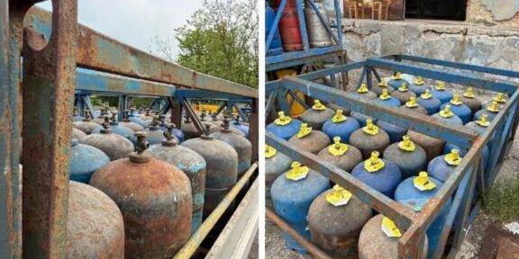 Rows of blue and rusty gas cylinders with yellow valve caps stored in a weathered metal rack outdoors.