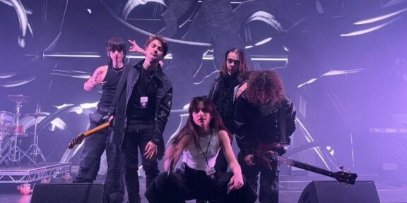 Five-member rock band poses on a stage bathed in purple lighting, with guitars and drums visible in the background.