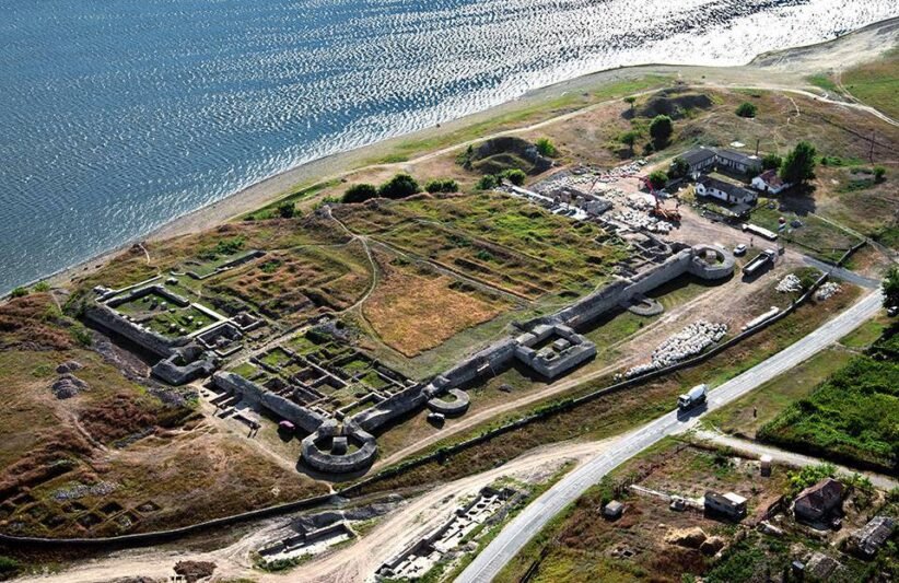 Aerial view of an old fortress with stone walls along a coastline, showing circular towers and grassy ruins near a road and small buildings nearby.